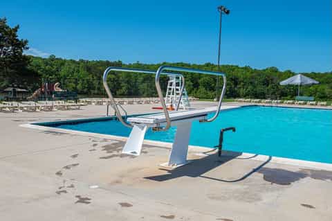 Resort swimming pool with diving board, lounge chairs, umbrellas, and wooded backdrop under clear blue sky