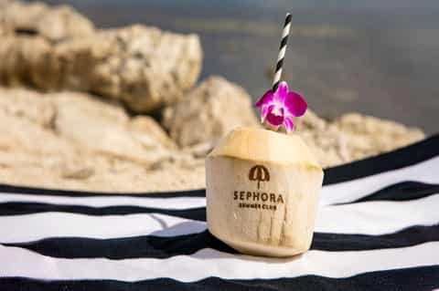 Wooden coconut cup with pink orchid and straw on striped towel by rocky beach at Sephora Summer Club