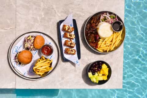 Overhead view of gourmet food spread by pool including burgers, steaks, and appetizers