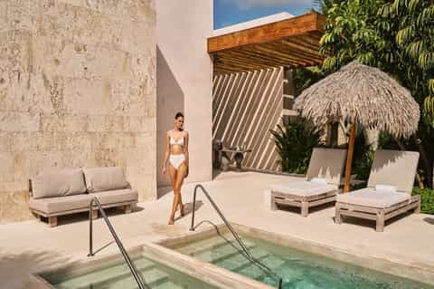 Woman in white bikini standing by geometric thermal pool with stone walls and thatched umbrellas