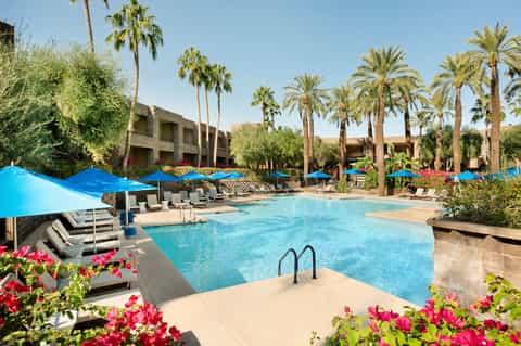 Resort pool with blue umbrellas, lounge chairs, palm trees, and desert architecture under clear sky