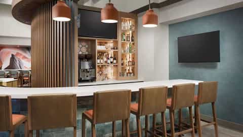 Modern hotel bar with copper pendant lights, white counter, wooden chairs with orange cushions, and teal accent wall with mounted TV