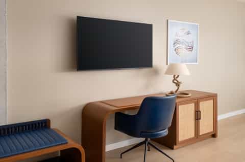 Hotel room workspace with mounted TV, wooden desk, navy chair, and nautical-themed wall art