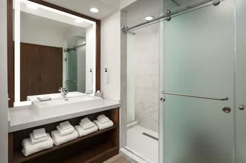 Modern hotel bathroom with illuminated vanity mirror, white sink, wooden accents, and walk-in shower