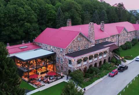 Stone luxury resort with pink metal roof, outdoor patio with red umbrellas, manicured grounds, and forest backdrop