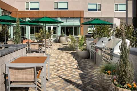 Patio dining area with green umbrellas, outdoor grills, and modern hotel building with herringbone pavers