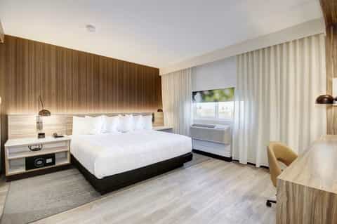 Modern hotel bedroom with queen bed, wooden headboard, light wood flooring, and white curtains framing windows