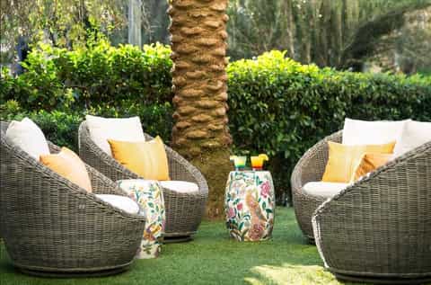 Two wicker lounge chairs with white and yellow pillows on grass beside palm tree and tropical greenery