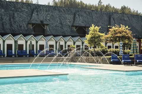 Swimming pool with water fountains and white beach cabanas backed by dark-roofed building