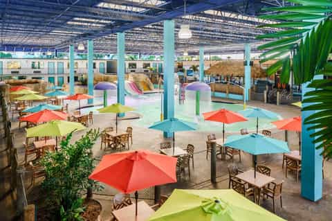Indoor water park facility with pools, colorful umbrellas, tropical plants, and wave pool