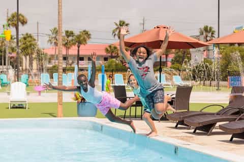 Kids jumping by pool edge with umbrellas, lounge chairs, and palm trees in background