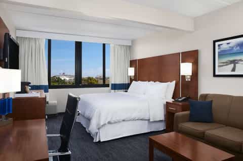 Upscale hotel room with queen bed, city skyline view, brown accent wall, and seating area