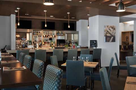 Modern restaurant interior with bar counter, teal patterned chairs, wooden tables, and pendant lighting fixtures
