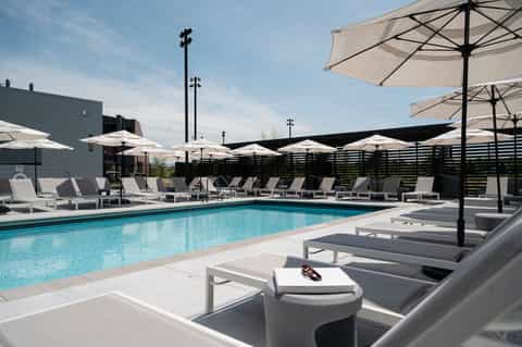 Contemporary pool area with white umbrellas, gray lounge chairs, and black privacy fence alongside building