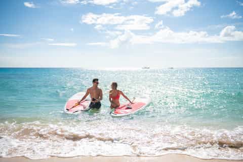 Couple with pink paddleboards wading in clear turquoise ocean water with white sandy beach