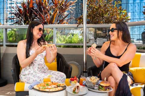 Two women dining outdoors on a rooftop terrace with city views, food and drinks on table, decorative plants in background