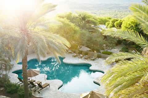 Aerial view of luxury resort pool surrounded by palm trees and desert landscape with mountains