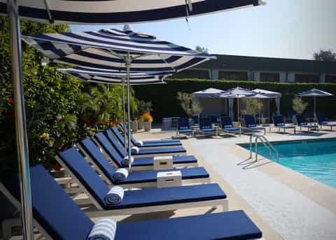 Poolside lounge area with striped umbrellas, blue and white loungers, and hedges under clear sky
