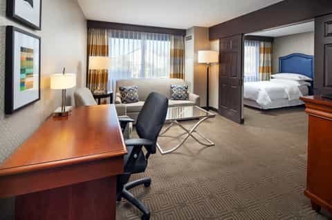 Spacious hotel suite with work desk, sitting area, and bedroom featuring warm wood furnishings and gold curtains