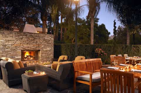 Outdoor fireplace lounge at dusk with stone walls, wooden furniture, and palm trees