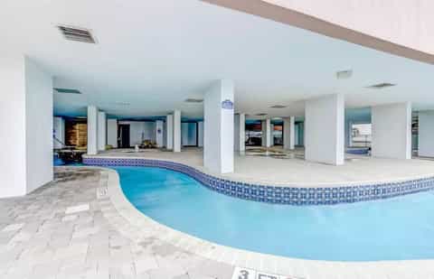 Indoor swimming pool with blue water and decorative tile border, white columns, and spacious facility with multiple entrances