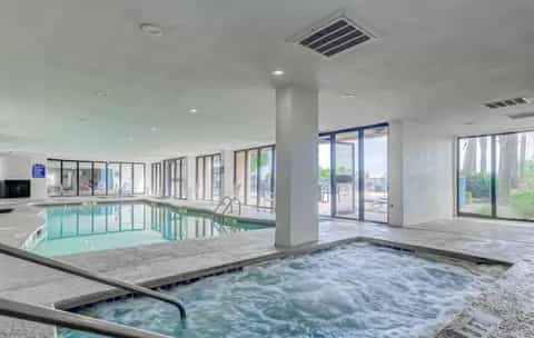 Indoor pool area with hot tub in foreground, large windows overlooking outdoor patio and landscaping, modern resort amenity space