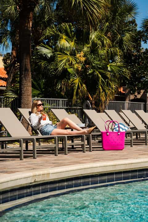 Woman relaxing on lounge chair by pool with palm trees, bright pink beach bag nearby