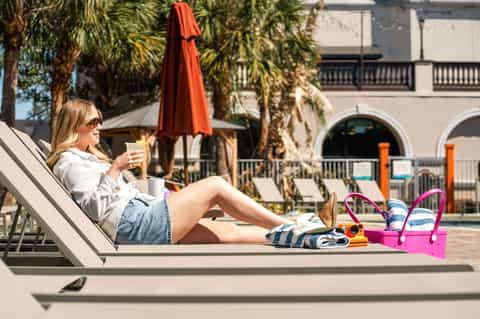 Woman lounging by resort pool with red umbrella, palm trees, and modern building architecture