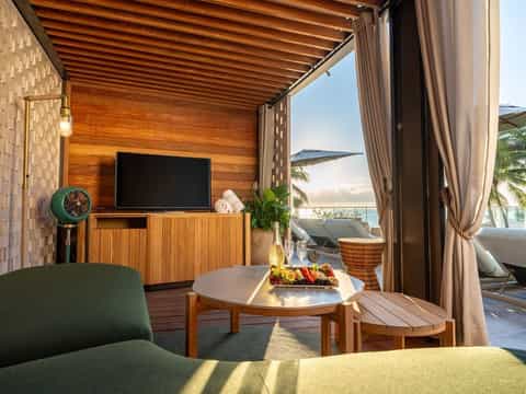Luxury resort cabana with wooden TV unit, ocean view through open curtains, green furniture, and beachside lounge visible