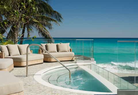 Beachfront infinity pool overlooking turquoise ocean with cream loungers, palm trees, and glass railing on pristine sand