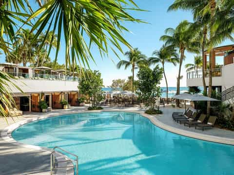 Curved resort pool with palm trees, ocean view, lounge chairs, and beachfront buildings under clear sky