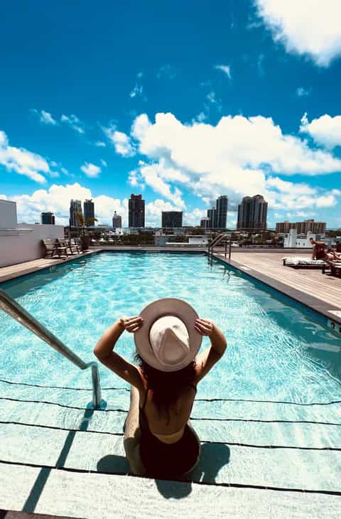 Person in hat at rooftop pool overlooking downtown city skyline under blue sky with white clouds