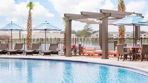 Resort pool deck with striped umbrellas, lounge chairs, palm trees, pergola, and water views