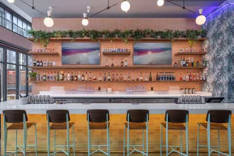 Modern bar with brick wall, wooden shelves displaying spirits, pendant lighting, and teal bar stools with yellow tile accents