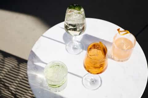 Cocktails on a white table including mojito, whiskey drink, and sparkling water in sunlight