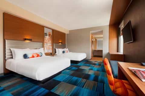 Double queen hotel room with wood headboards, white linens, blue patterned carpet, and work desk
