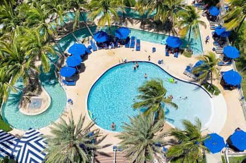 Aerial view of resort pool with blue umbrellas, palm trees, and adjacent lazy river surrounded by lounge chairs