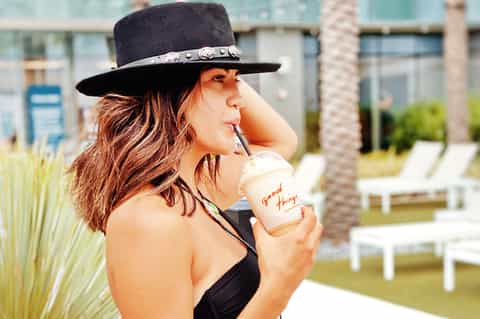Woman in black bikini wearing a black hat, holding a beverage at a resort poolside with lounge chairs