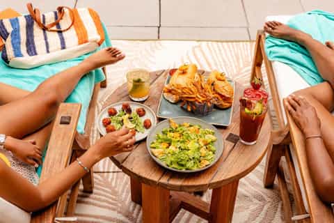 Two people enjoying lunch at a wooden table with salads, fries, and beverages on a terrace