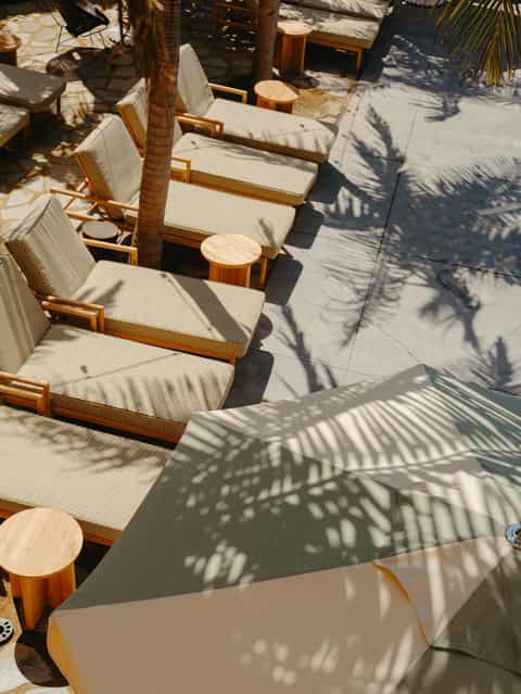 Overhead view of wooden lounge chairs with cream cushions arranged in a row on a sunny poolside patio with palm tree shadows