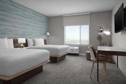 Modern hotel room with two queen beds, blue accent wall, desk, TV, and large window with city views