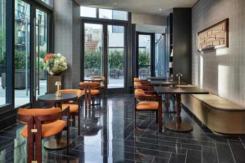 Modern hotel lobby with black marble floors, orange chairs, dark wood tables, tiled walls, and glass doors leading to terrace