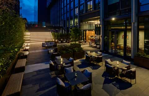 Nighttime courtyard with illuminated steps, lounge seating, planters, and lit building facade creating contemporary outdoor space
