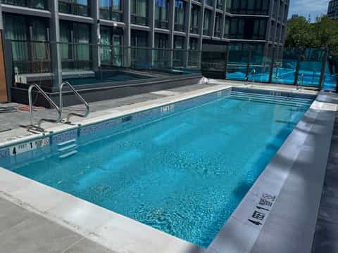 Indoor swimming pool with turquoise water, white deck, metal railings, glass barriers, and high-rise windows in background