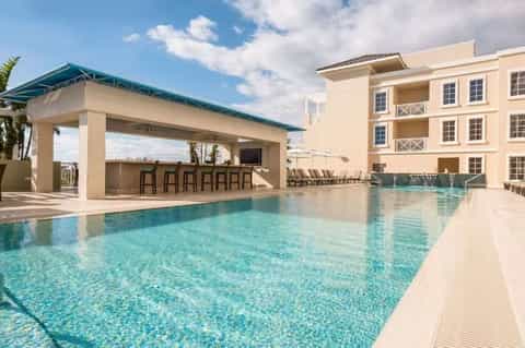 Resort pool with modern poolside bar pavilion, lounge chairs, and cream-colored building with tropical landscaping