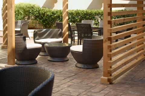 Patio seating area with woven chairs, white cushions, wooden pergola, and hedged privacy screening