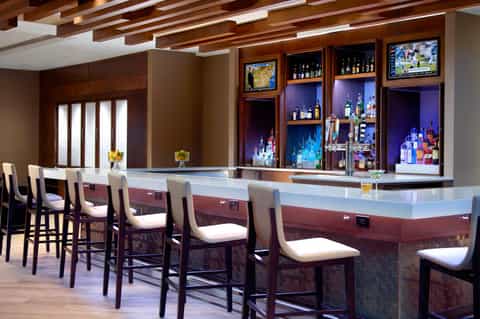 Upscale bar lounge with white counter, wooden ceiling beams, illuminated liquor shelves, and cushioned bar stools