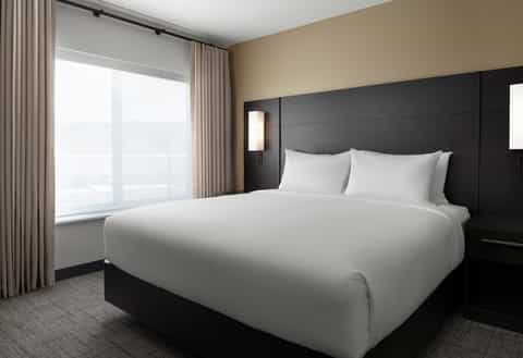 Modern hotel bedroom with king bed, black headboard, beige walls, and floor-to-ceiling windows with city views
