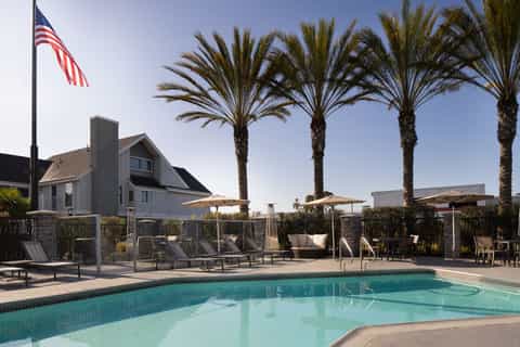 Coastal pool area with palm trees, umbrella shade, lounge chairs, and modern cottage-style building