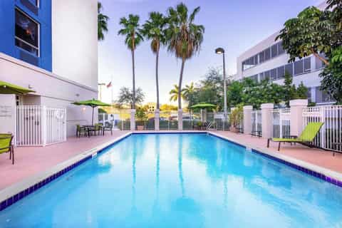 Resort outdoor swimming pool with palm trees, white fence, green umbrellas, and pink surrounding patio area